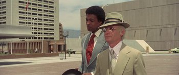 Movie still from “The Man Who Fell to Earth” (1976), directed by Nicolas Roeg – A man in a suit and a man in a hat and sunglasses; Medium shot, Low angle