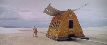Movie still from “The Man Who Fell to Earth” (1976), directed by Nicolas Roeg – Two people standing in front of a building on the beach; Extreme Wide shot, Low angle