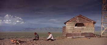 Movie still from “The Man Who Fell to Earth” (1976), directed by Nicolas Roeg – A man and a woman sitting in the sand near a shack; Wide shot, Low angle