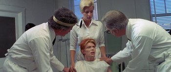 Movie still from “The Man Who Fell to Earth” (1976), directed by Nicolas Roeg – A group of people standing next to a man in a room; Medium shot, Over the shoulder angle