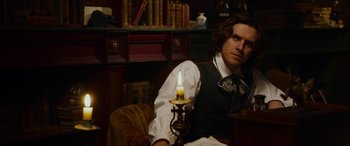 Movie still from “The Man Who Invented Christmas” (2017), directed by Bharat Nalluri – A man sitting in a chair holding a lit candle; Medium shot, Over the shoulder angle