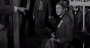 Movie still from “The Man Who Shot Liberty Valance” (1962), directed by John Ford – A man in a suit and tie sitting in front of a group of men; Medium shot, Over the shoulder angle