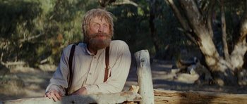 Movie still from “The Man from Snowy River” (1982), directed by George Miller – An old man with a beard sitting on top of a wooden bench; Medium shot, Low angle