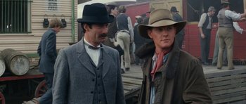 Movie still from “The Man from Snowy River” (1982), directed by George Miller – Two men in hats and suits standing next to each other on a train platform; Medium shot, Over the shoulder angle