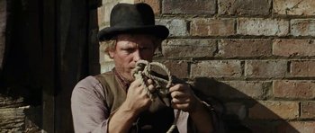 Movie still from “The Man from Snowy River” (1982), directed by George Miller – A man wearing a hat and holding a rope in front of a brick wall; Close Up shot, Low angle