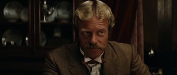 Movie still from “The Man from Snowy River” (1982), directed by George Miller – An older man with a mustache and a beard wearing a suit and tie; Close Up shot, Over the shoulder angle