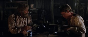 Movie still from “The Man from Snowy River” (1982), directed by George Miller – Two people sitting at a table in a dimly - lit room; Medium shot, Over the shoulder angle