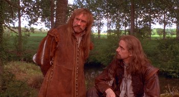 Movie still from “The Man in the Iron Mask” (1998), directed by Randall Wallace – Two men dressed in medieval clothing sitting next to each other in a wooded area; Medium shot, Over the shoulder angle