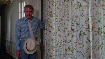 Movie still from “The Man with Two Brains” (1983), directed by Carl Reiner – A man wearing a hat and a tie standing in front of a curtain; Medium shot, Low angle