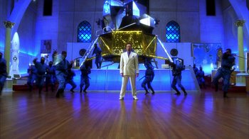 Movie still from “The Master of Disguise” (2002), directed by Perry Andelin Blake – A man in a white suit standing in front of a large sculpture; Wide shot, High angle