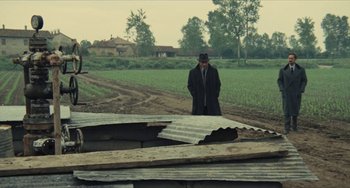 Movie still from “The Mattei Affair” (1972), directed by Francesco Rosi – An old man in a black coat and a black hat is standing in front of a field; Wide shot, High angle