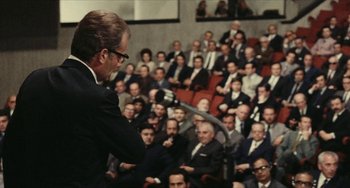 Movie still from “The Mattei Affair” (1972), directed by Francesco Rosi – A man in a black suit is holding a microphone in front of an audience; Wide shot, Over the shoulder angle