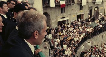 Movie still from “The Mattei Affair” (1972), directed by Francesco Rosi – An older man wearing a green tie speaking in front of a large crowd of onlookers; Extreme Wide shot, High angle