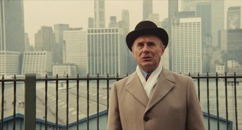 Movie still from “The Mattei Affair” (1972), directed by Francesco Rosi – An older man in a suit and hat standing in front of a city; Medium shot, Low angle