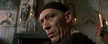 Movie still from “The Messenger: The Story of Joan of Arc” (1999), directed by Luc Besson – An older man wearing a black hat looking to his left; Close Up shot, Low angle