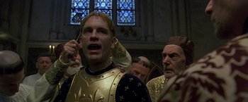Movie still from “The Messenger: The Story of Joan of Arc” (1999), directed by Luc Besson – A man in a gold and black suit is talking on a cell phone; Close Up shot, Low angle