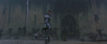 Movie still from “The Messenger: The Story of Joan of Arc” (1999), directed by Luc Besson – A man in armor holding a sword in front of a wall; Wide shot, Low angle