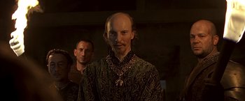 Movie still from “The Messenger: The Story of Joan of Arc” (1999), directed by Luc Besson – A man with a bald head wearing a necklace; Close Up shot, Over the shoulder angle