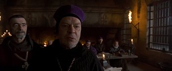 Movie still from “The Messenger: The Story of Joan of Arc” (1999), directed by Luc Besson – An old man wearing a purple hat in front of a group of people; Close Up shot, Over the shoulder angle