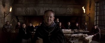 Movie still from “The Messenger: The Story of Joan of Arc” (1999), directed by Luc Besson – A group of people sitting in a room with a priest; Medium shot, Over the shoulder angle