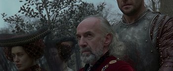 Movie still from “The Messenger: The Story of Joan of Arc” (1999), directed by Luc Besson – An older man with long white hair wearing a red coat; Close Up shot, Over the shoulder angle