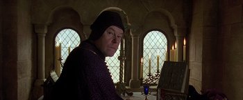 Movie still from “The Messenger: The Story of Joan of Arc” (1999), directed by Luc Besson – A man wearing a black hat in front of stained glass windows; Medium shot, Low angle