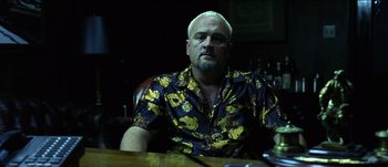 Movie still from “The Mexican” (2001), directed by Gore Verbinski – A man sitting at a wooden table wearing a yellow shirt; Medium shot, Low angle