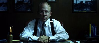 Movie still from “The Mexican” (2001), directed by Gore Verbinski – A man sitting at a table with his arms crossed; Medium shot, Over the shoulder angle