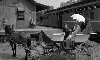 Movie still from “The Miracle Worker” (1962), directed by Arthur Penn – A man and a woman sitting on a horse drawn carriage in front of a train station; Wide shot, Low angle