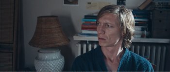 Movie still from “The Misfortunates” (2009), directed by Felix van Groeningen – A man sitting in front of a stack of books in a room; Close Up shot, Over the shoulder angle