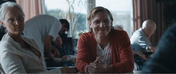 Movie still from “The Misfortunates” (2009), directed by Felix van Groeningen – A man sitting at a table smiling for the camera; Medium shot, Over the shoulder angle