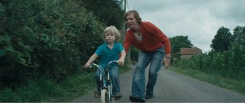 Movie still from “The Misfortunates” (2009), directed by Felix van Groeningen – A woman and a boy riding a bike down a road; Medium shot, Low angle