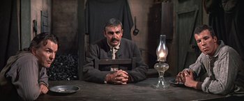 Movie still from “The Molly Maguires” (1970), directed by Martin Ritt – A man sitting at a table in a room with an oil lamp; Medium shot, Over the shoulder angle