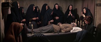 Movie still from “The Molly Maguires” (1970), directed by Martin Ritt – A group of people sitting around a man laying on a bed; Wide shot, High angle
