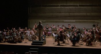 Movie still from “The Money Pit” (1986), directed by Richard Benjamin – A group of people sitting on a stage with a conductor; Extreme Wide shot, High angle
