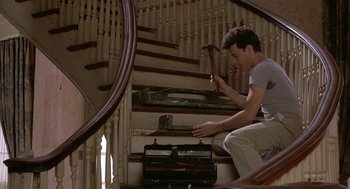 Movie still from “The Money Pit” (1986), directed by Richard Benjamin – A man sitting on the bottom of a set of stairs holding a hammer; Medium shot, Low angle