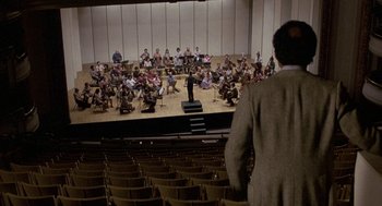 Movie still from “The Money Pit” (1986), directed by Richard Benjamin – A man standing in front of an audience in front of an orchestra; Extreme Wide shot, Over the shoulder angle
