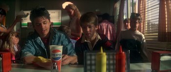 Movie still from “The Monster Squad” (1987), directed by Fred Dekker – A young man sitting at a table in front of two other people; Medium shot, Over the shoulder angle