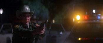 Movie still from “The Monster Squad” (1987), directed by Fred Dekker – A man holding a gun while standing in front of a car; Medium shot, Over the shoulder angle