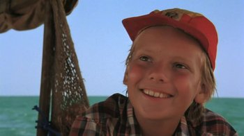 Movie still from “The Mosquito Coast” (1986), directed by Peter Weir – A young boy wearing a hat smiling for the camera; Close Up shot, Low angle