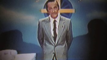 Movie still from “The Mosquito Coast” (1986), directed by Peter Weir – A man in a suit and tie standing in front of a clock; Medium shot, High angle