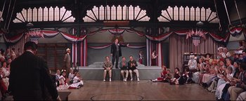 Movie still from “The Music Man” (1962), directed by Morton DaCosta – A group of people sitting on a stage in front of an audience; Wide shot, Low angle