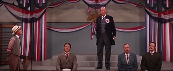 Movie still from “The Music Man” (1962), directed by Morton DaCosta – A man in a suit and tie standing on a stage next to another man in a suit and tie; Medium shot, Low angle