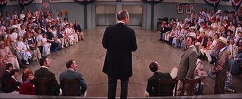Movie still from “The Music Man” (1962), directed by Morton DaCosta – A group of people sitting in a circle around a man in a black suit; Wide shot, High angle