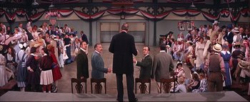 Movie still from “The Music Man” (1962), directed by Morton DaCosta – A group of men standing around each other in front of a crowd; Wide shot, High angle