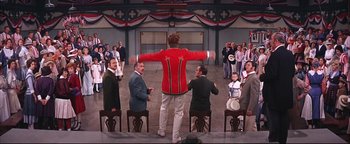 Movie still from “The Music Man” (1962), directed by Morton DaCosta – A group of people standing around a man in a red jacket; Wide shot, High angle