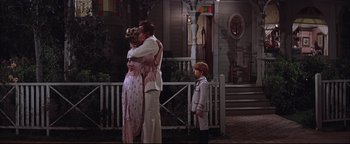Movie still from “The Music Man” (1962), directed by Morton DaCosta – Two people hug in front of a house while two children look on; Wide shot, Over the shoulder angle
