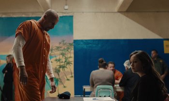 Movie still from “The Mustang” (2019), directed by Laure de Clermont-Tonnerre – A man in an orange jumpsuit standing next to a table; Medium shot, Over the shoulder angle