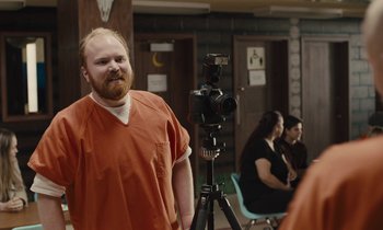 Movie still from “The Mustang” (2019), directed by Laure de Clermont-Tonnerre – A man in an orange shirt standing in front of a camera; Medium shot, Over the shoulder angle