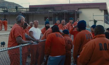 Movie still from “The Mustang” (2019), directed by Laure de Clermont-Tonnerre – A group of men in orange jumpsuits gathered around a chain link fence; Wide shot, High angle